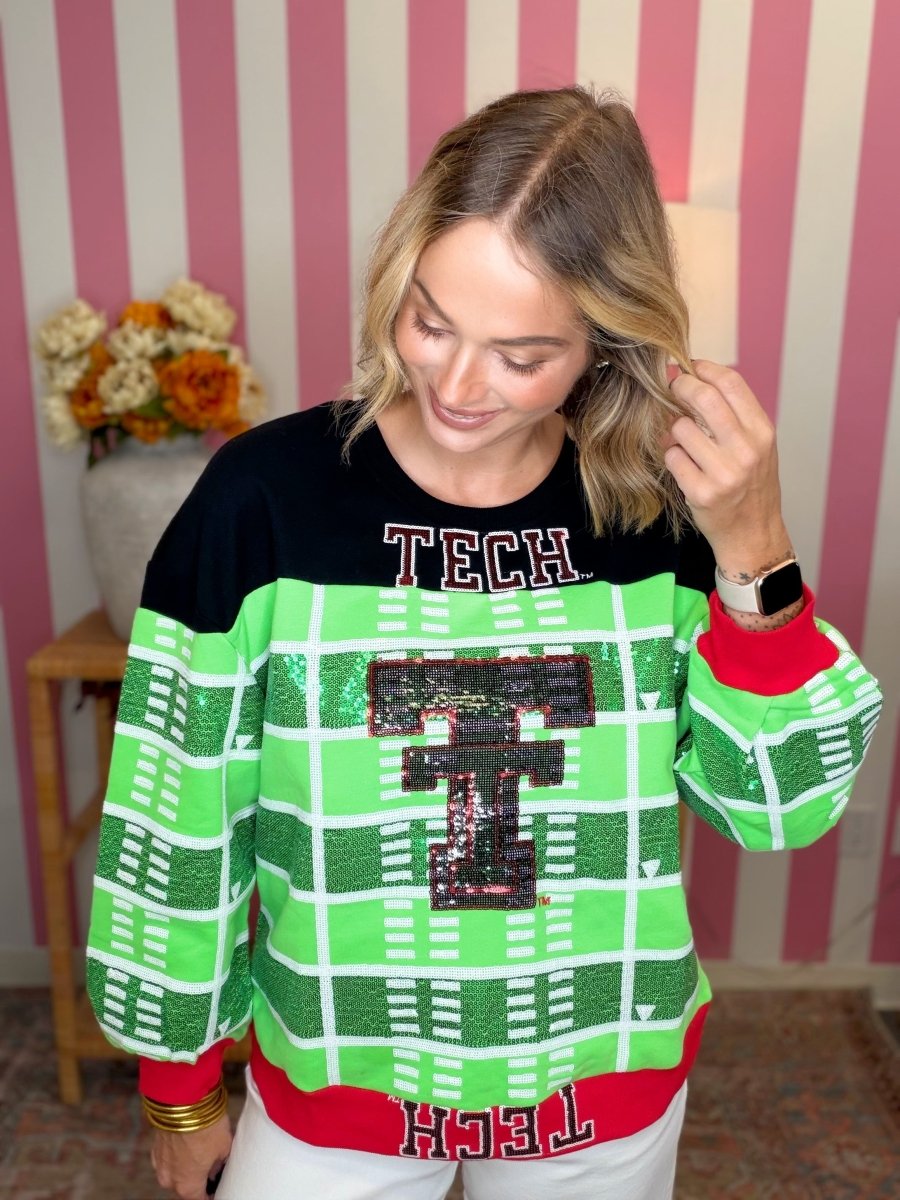 A woman wearing a festive 'Tech' themed sweater with a black upper section, green and white football field plaid pattern, and red accents, posed against a pink and white striped background.