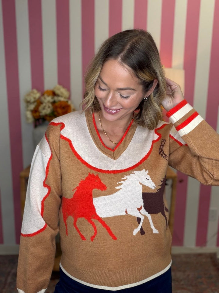A woman wearing a brown knit sweater with a cream V-neck collar, red and white striped cuffs, and a graphic of red, white, and brown horses on the front, posed in front of pink and white striped wallpaper.
