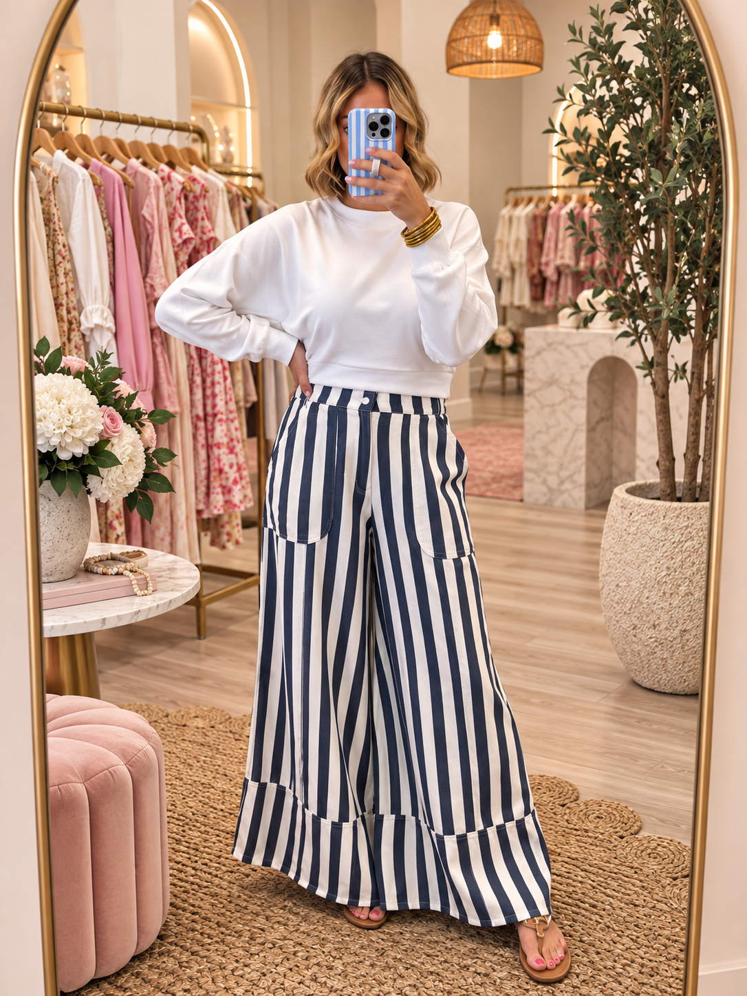A woman takes a mirror selfie in a boutique wearing a white long-sleeve mock neck top and navy and white vertically striped wide-leg pants. The boutique features pink clothing racks, a large gold-framed mirror, and a cozy interior with a pink velvet ottoman and a potted tree.