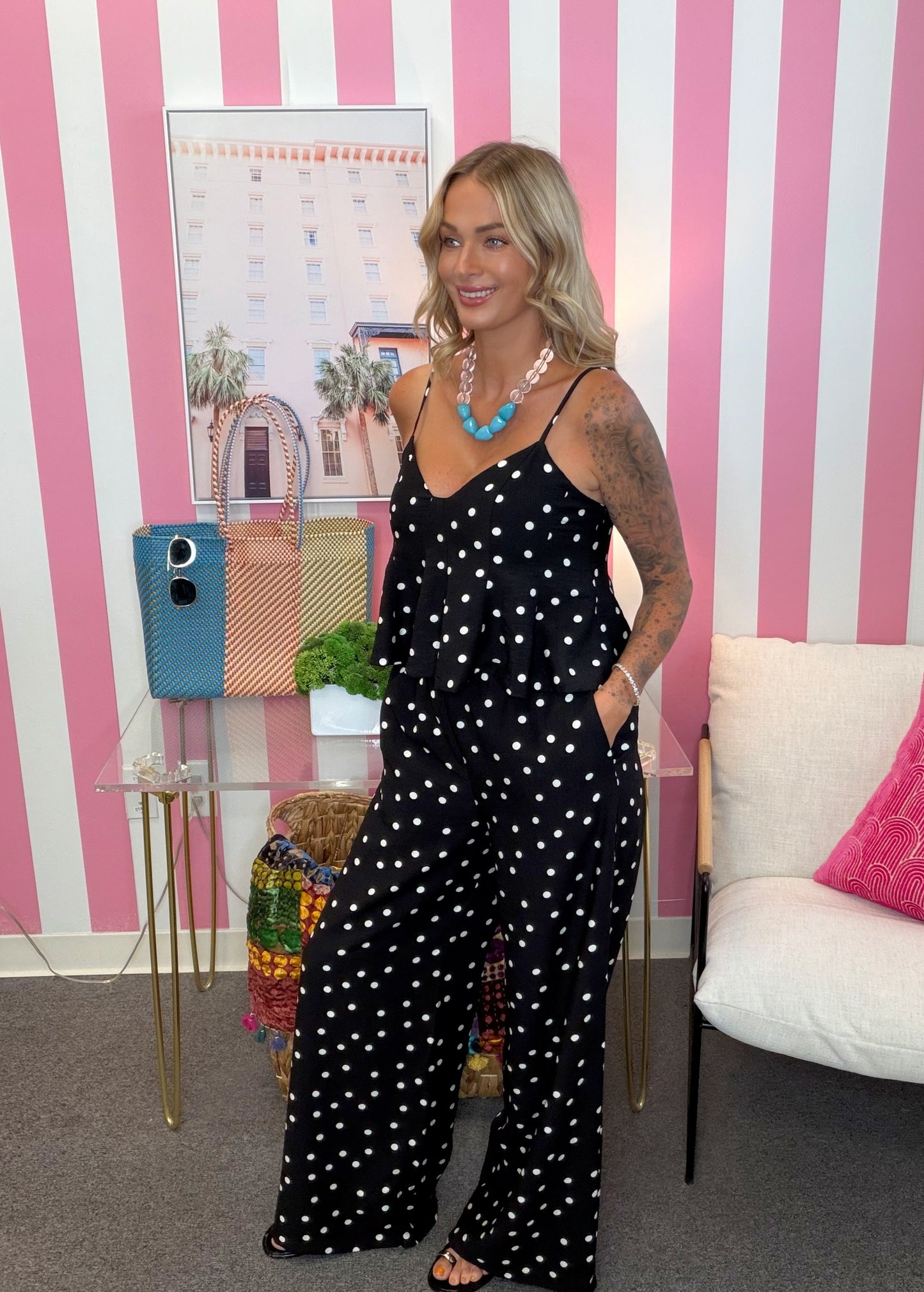 A woman stands in a pink and white striped room wearing a black sleeveless jumpsuit with white polka dots. She is accessorized with a chunky turquoise necklace and a pearl necklace, standing next to a small glass table with colorful handbags and a white armchair.