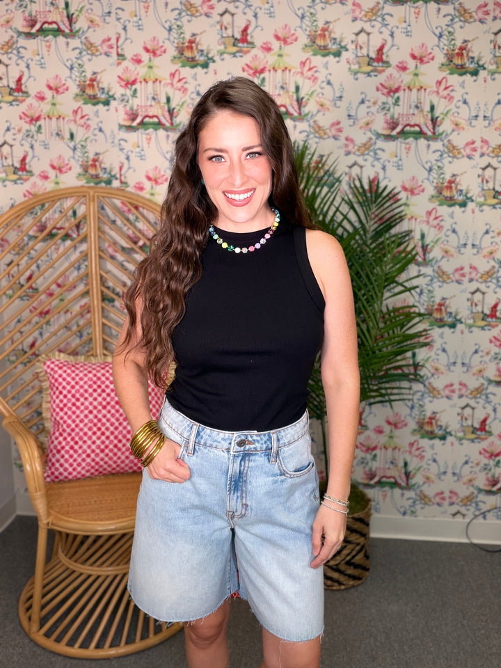 Woman wearing a black sleeveless top and denim shorts standing in a room with floral wallpaper.