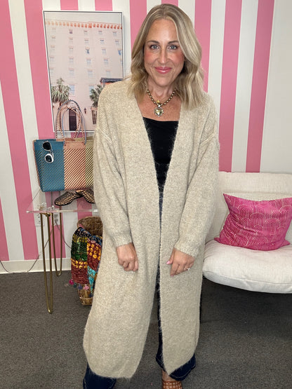 A woman wearing a long, open-front, oversized knit cardigan in a light oatmeal beige color, styled over a black top. She is standing in a room with pink and white striped walls, a white sofa with a pink pillow, and a small side table.