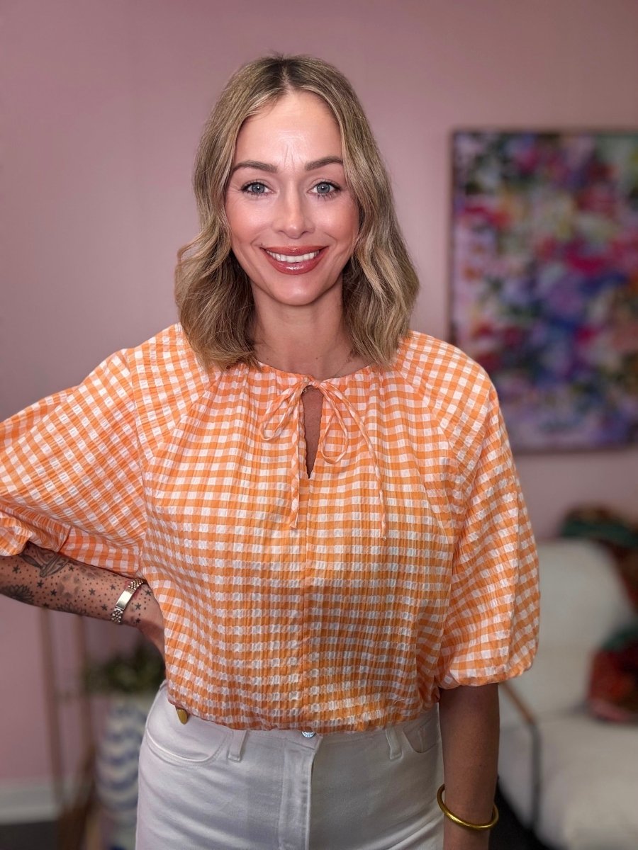 Woman wearing an orange checkered blouse in a room with a colorful painting on the wall.