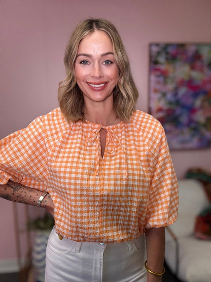 Woman wearing an orange checkered blouse in a room with a colorful painting on the wall.