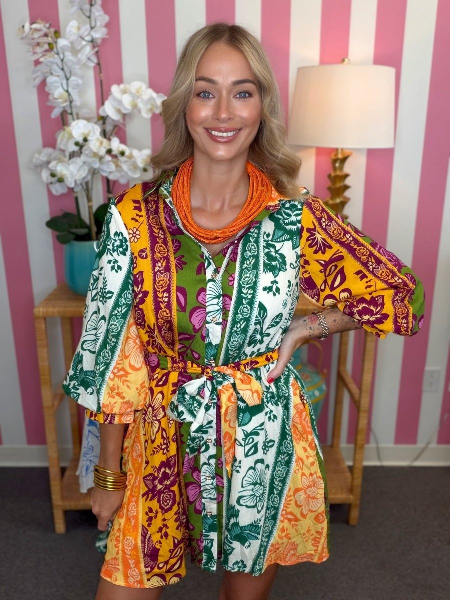 A woman modeling a colorful, patterned mini dress with wide three-quarter sleeves and a tie waist, styled with a chunky orange necklace, standing in front of a pink and white striped wall.