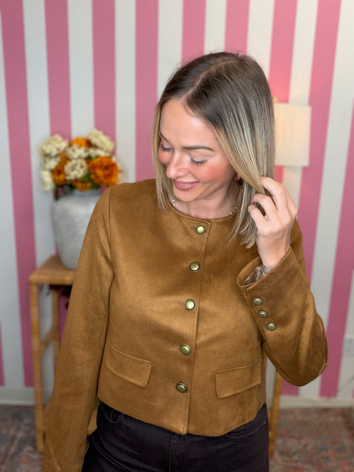 A woman wearing a cropped brown suede jacket with gold buttons, standing in front of a pink and white striped wall.