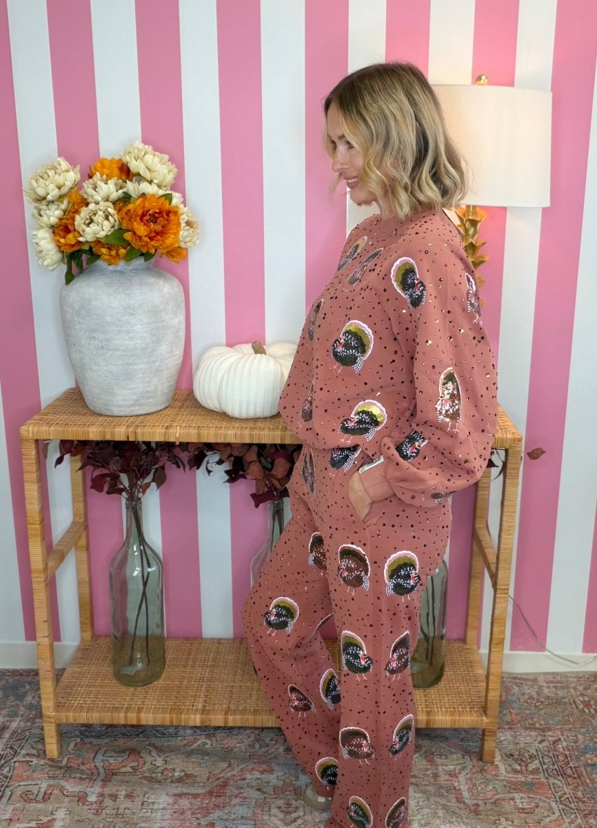 A woman wearing a dusty rose pajama set with a black and white skull pattern stands in profile next to a wooden console table decorated with a vase of flowers, a white pumpkin, and glass bottles, against a pink and white striped wall.