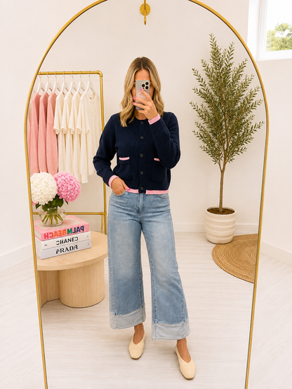 A woman modeling a dark navy blue cardigan with pink pocket trim and a pink hem, paired with light-wash blue jeans and cream-colored shoes, standing in front of a pink and white striped wall.