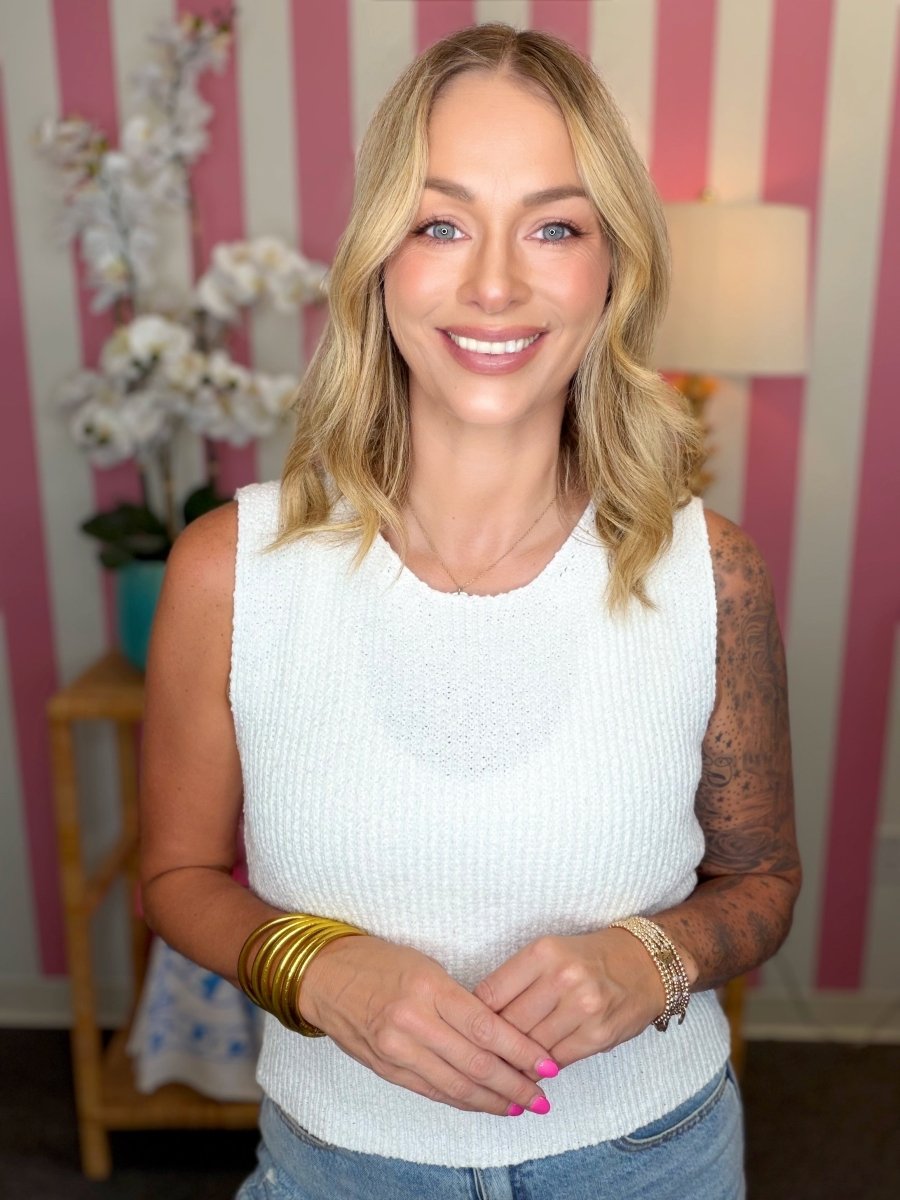 A smiling blonde woman wearing a white ribbed sleeveless top and blue jeans, standing indoors against pink and white striped wallpaper with a lamp and flowers in the background.