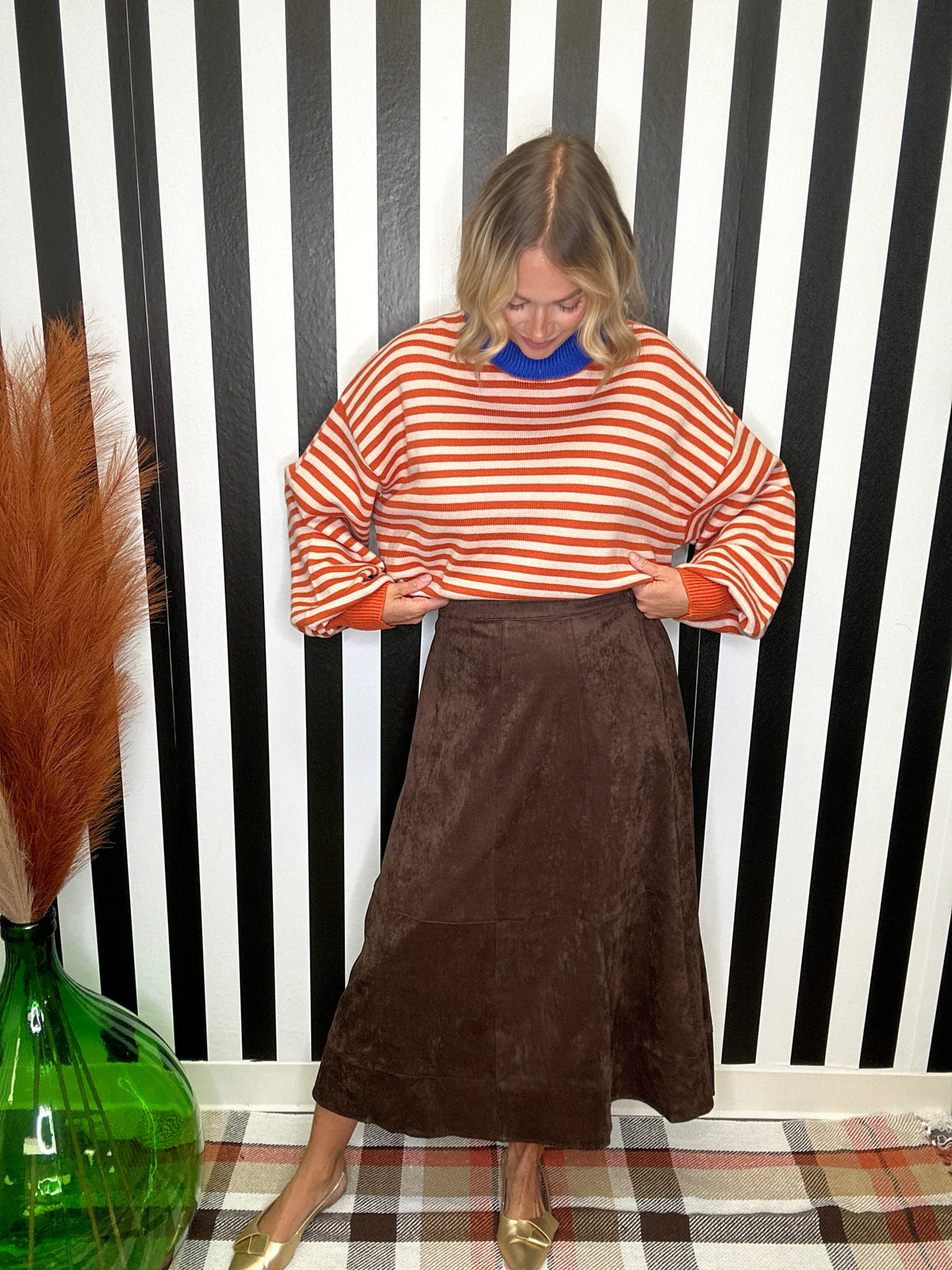 A woman modeling a rust-orange and white striped knit sweater with a blue crew neck, paired with a brown corduroy skirt, standing in front of a black and white striped wall.