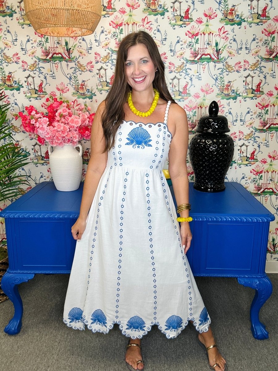Woman in a white dress with blue patterns standing in front of a colorful wall with floral designs.