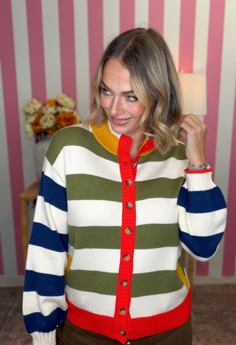 A woman wearing a colorful striped cardigan sweater with green, white, blue, and red horizontal stripes, standing in front of a pink and white striped wall.