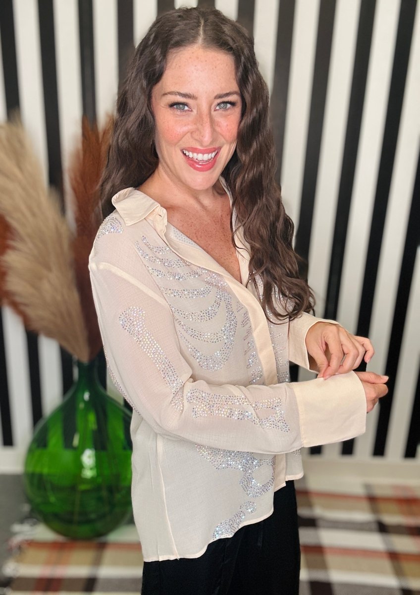 A woman with long wavy brown hair smiling while wearing a sheer cream button-up blouse with silver sequin embroidery, paired with black pants. She is standing in front of a black and white striped wall with a green glass vase and pampas grass in the background.