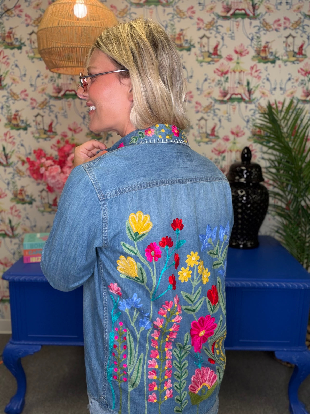 A woman wearing a light blue denim jacket with colorful floral embroidery on the back, standing in front of floral wallpaper and a blue side table.