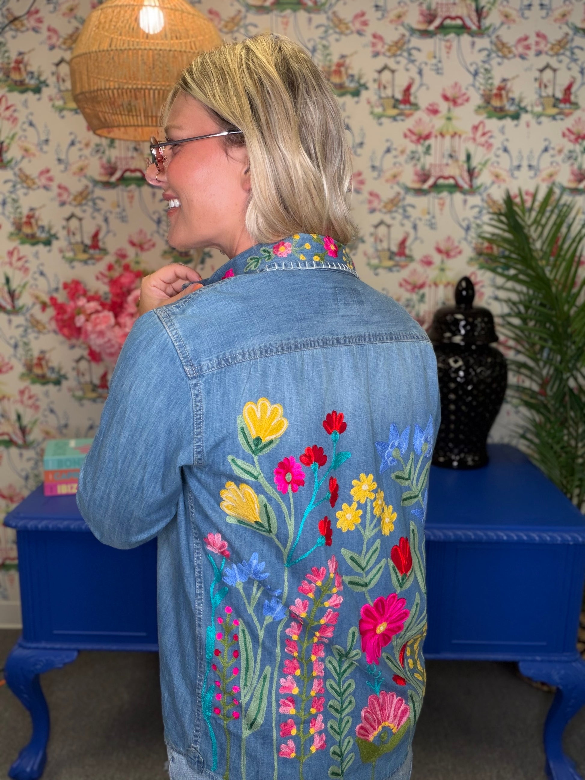 A woman wearing a light blue denim jacket with colorful floral embroidery on the back, standing in front of floral wallpaper and a blue side table.