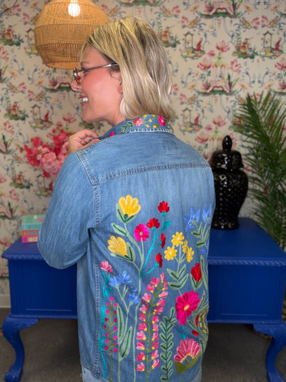 A woman wearing a light blue denim jacket with colorful floral embroidery on the back, standing in front of floral wallpaper and a blue side table.