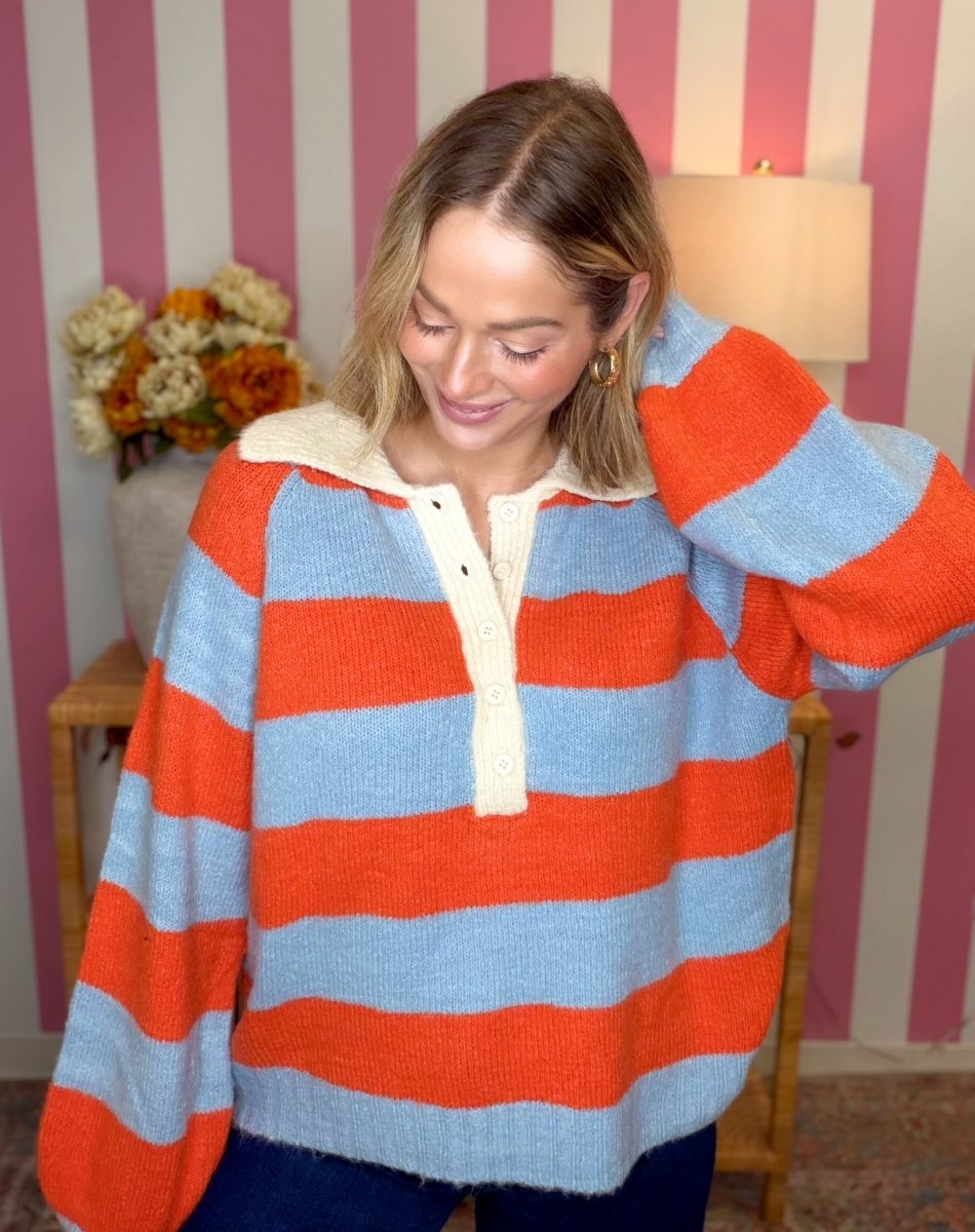 A woman wearing a fuzzy knit sweater with bold horizontal stripes in orange and light blue, featuring a cream-colored collar and button placket. She is standing in front of a pink and white striped wall.
