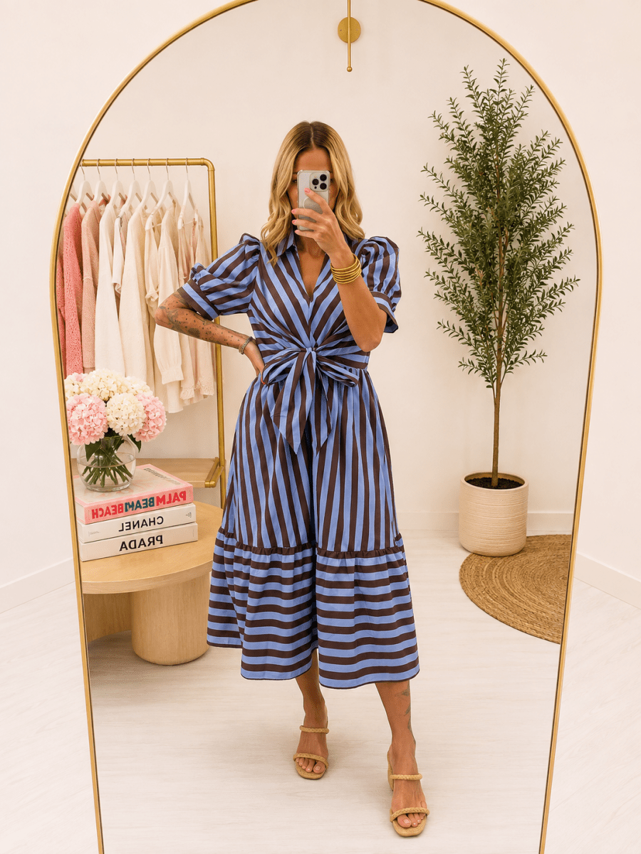 A woman takes a mirror selfie in a boutique wearing a blue and brown vertically striped midi dress with puff sleeves and a tie waist. The boutique features a gold-framed arch mirror, a clothing rack with neutral-colored dresses, a small table with flowers, and a potted plant.
