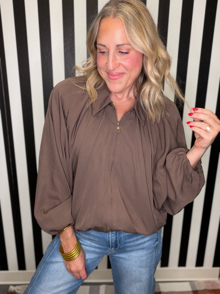 A woman wearing a brown long-sleeve blouse with a front zipper and blue jeans, standing in front of a black and white striped wall.