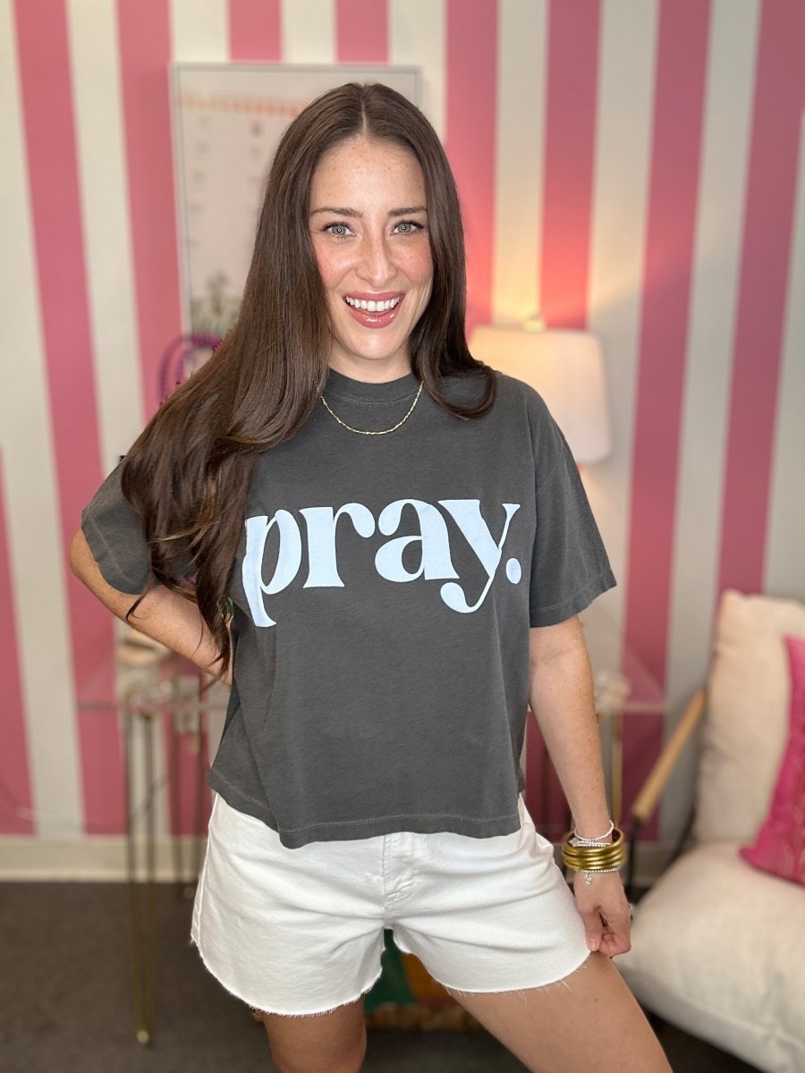 Woman wearing a gray t-shirt with 'pray.' printed on it, standing in a room with striped walls.