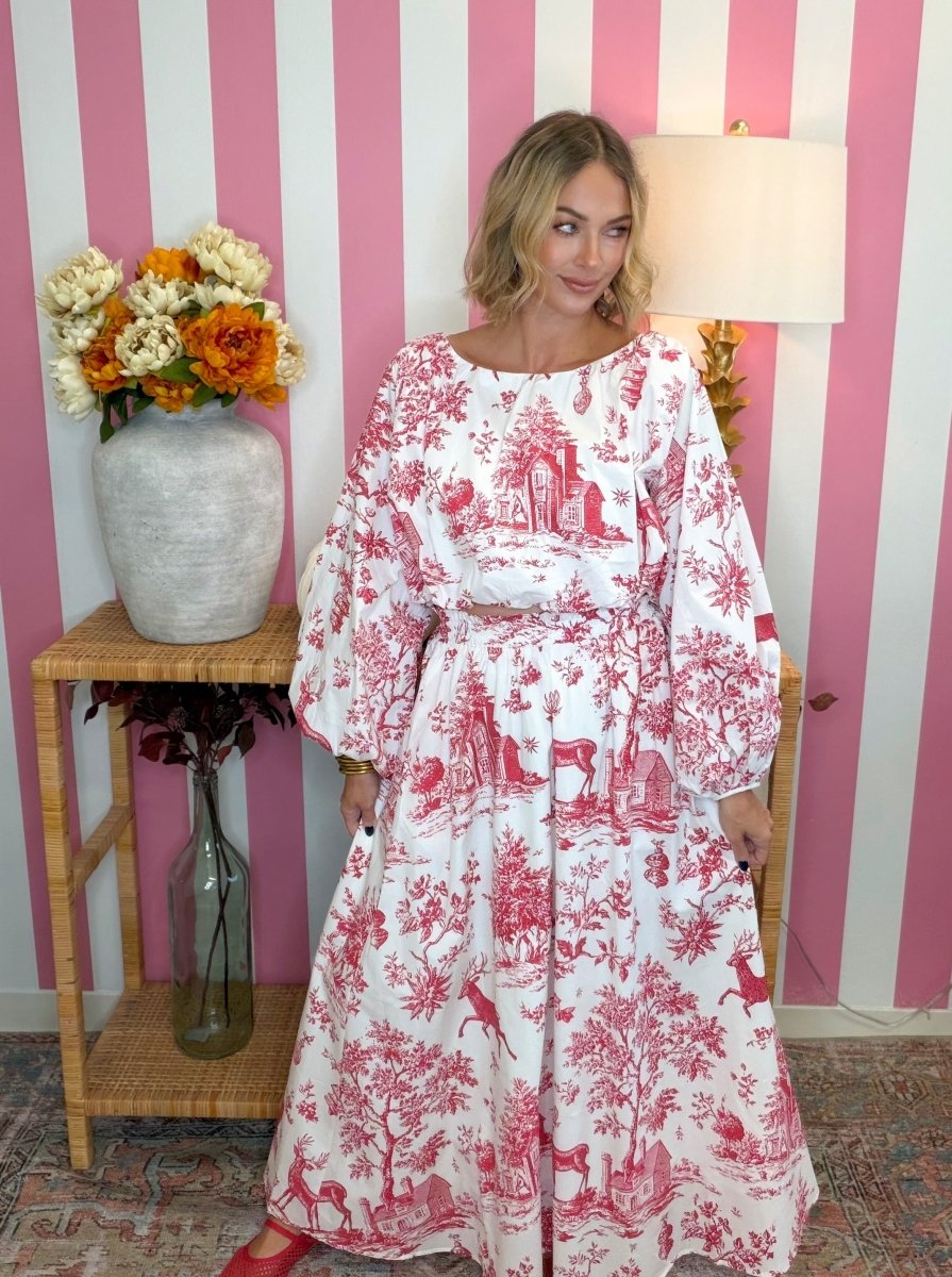 A woman wearing a long-sleeved white maxi dress with a red toile de Jouy print stands in front of a pink and white striped wall. She is posing next to a wooden side table with a vase of flowers and a lamp.