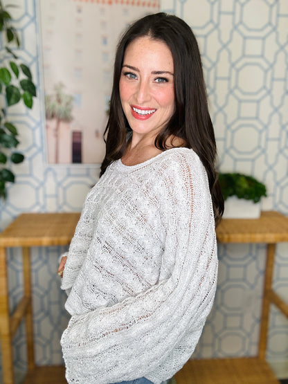 Woman wearing a light gray sweater in a room with patterned wallpaper and wooden furniture.