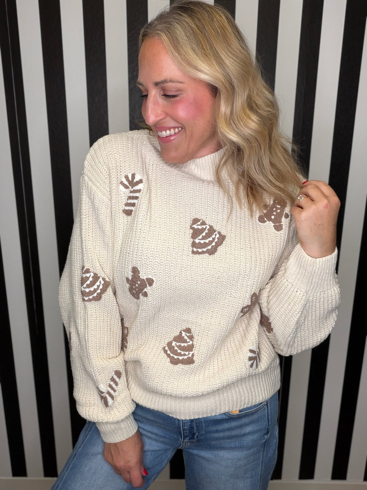 A woman wearing a cream-colored knit sweater with brown gingerbread cookie motifs, paired with blue jeans, standing in front of a black and white striped background.