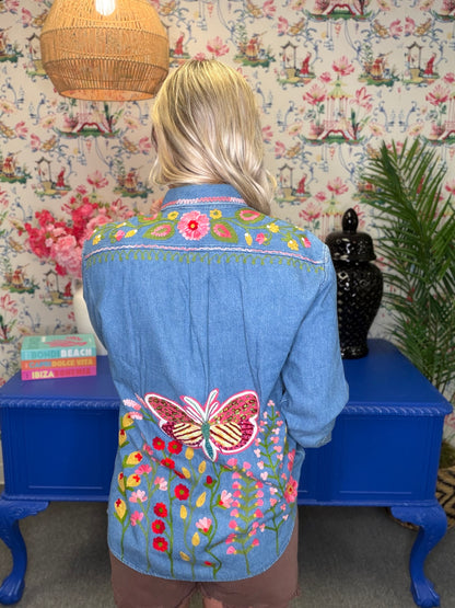 A woman viewed from behind wearing a light blue denim jacket decorated with colorful floral and butterfly embroidery, standing in front of a blue table and floral wallpaper.