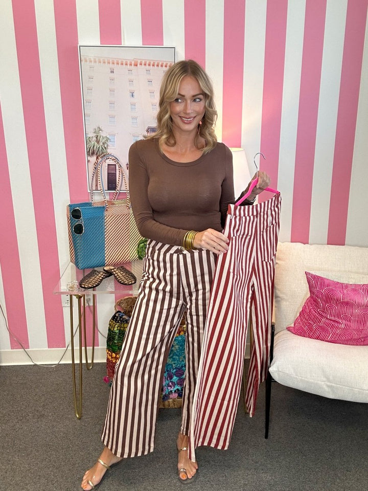 A woman stands in a room with pink and white striped wallpaper, wearing brown wide-leg striped pants and holding up matching striped pants on a hanger. She is also wearing a brown long-sleeve top and beige heels.