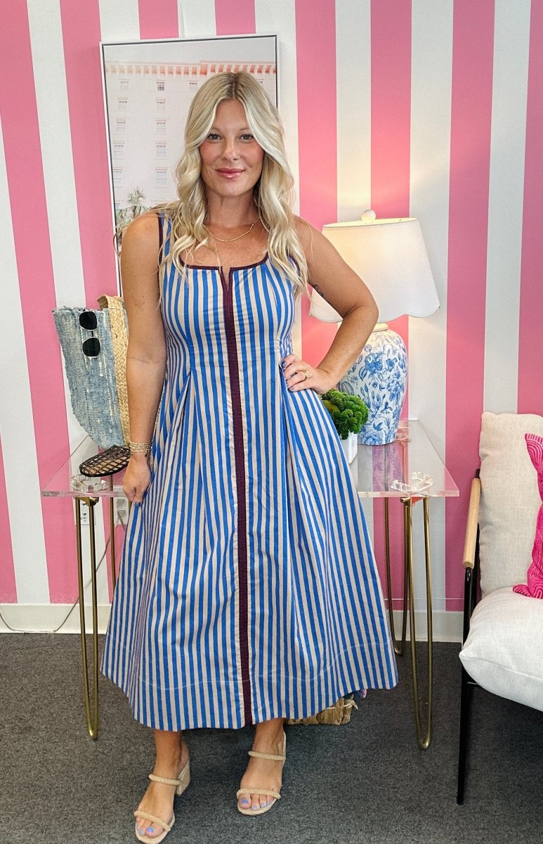 A woman modeling a sleeveless blue and white vertically striped midi dress with a front zipper, standing in front of a pink and white striped wall.
