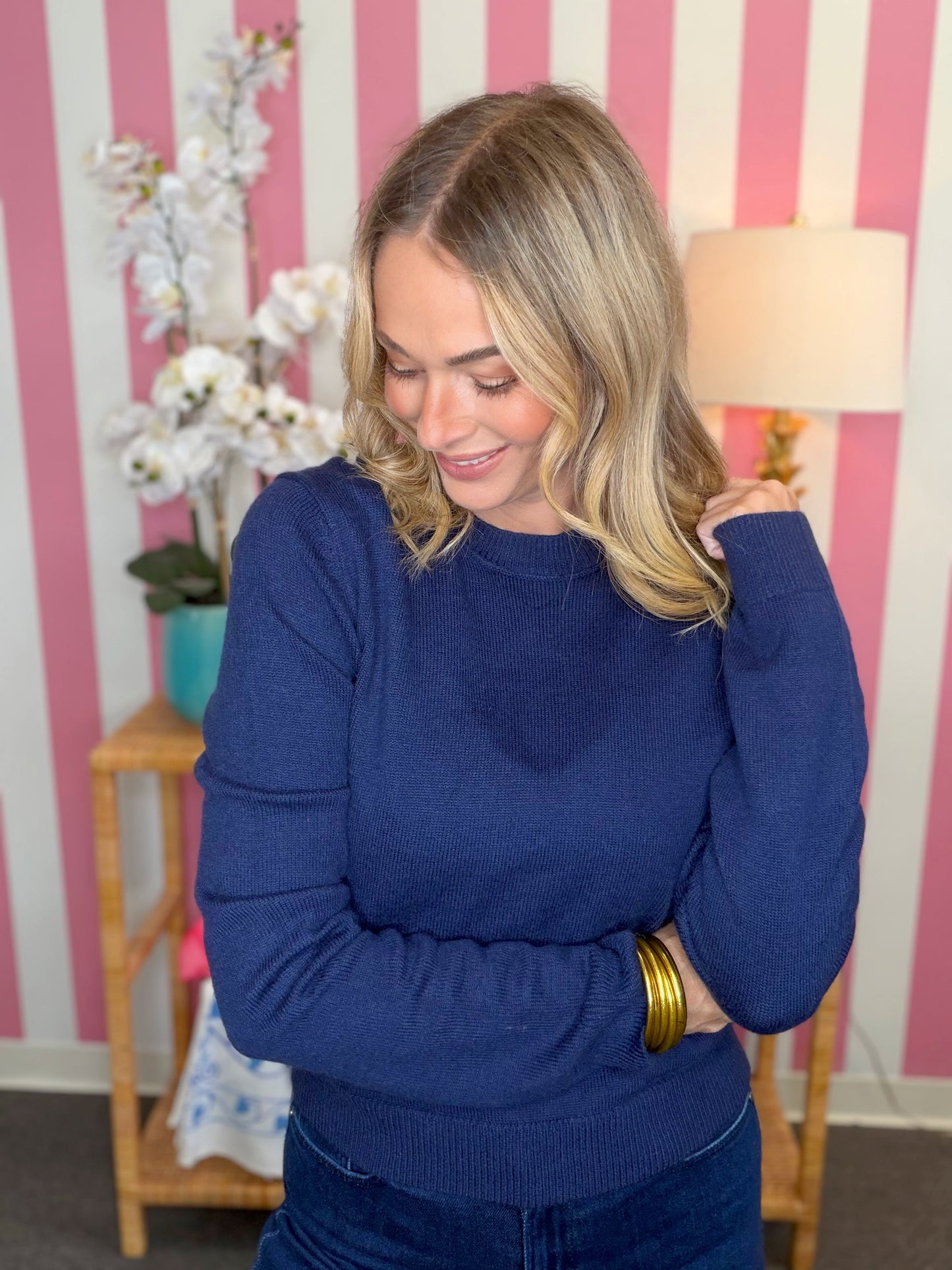 A woman with blonde hair wearing a navy blue knit sweater and dark blue jeans, standing in front of a pink and white striped wall with a lamp and flowers in the background.