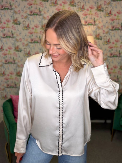 A woman wearing a white satin button-up blouse with black scalloped trim on the collar and front placket, paired with blue jeans, standing in front of a floral wallpaper background.