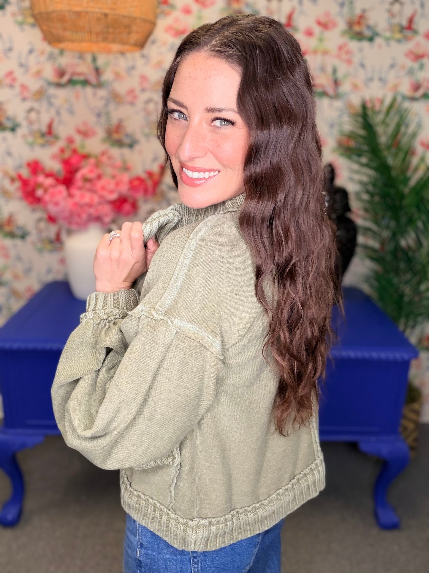 A woman with long wavy brown hair is shown from the back wearing an olive green knit sweater with ruffled trim, standing in a decorated room with floral wallpaper, a blue bench, and plants.