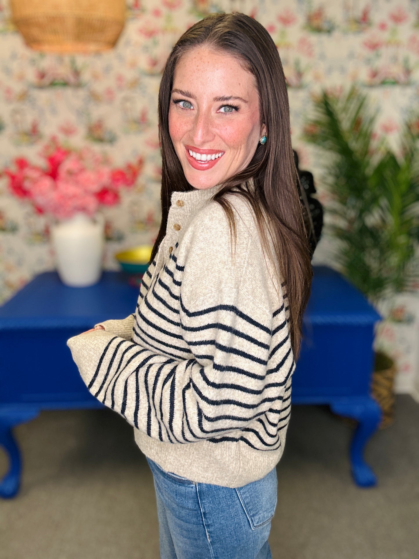A woman with long brown hair smiling and looking over her shoulder, wearing a cream knit sweater with black wavy stripes and blue jeans, standing in a decorated room with a blue table and floral wallpaper.