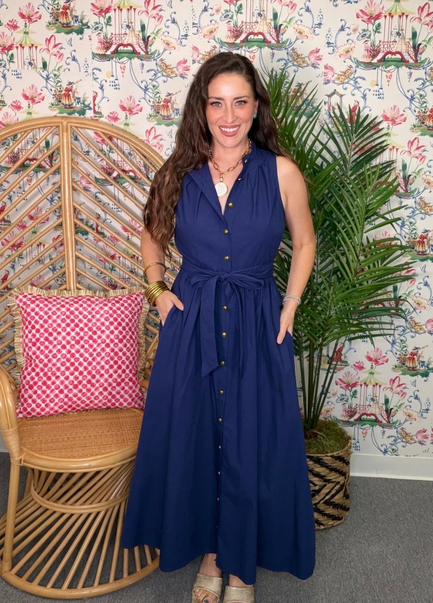 Woman in a blue dress standing in a room with floral wallpaper, a wicker chair, and a plant.