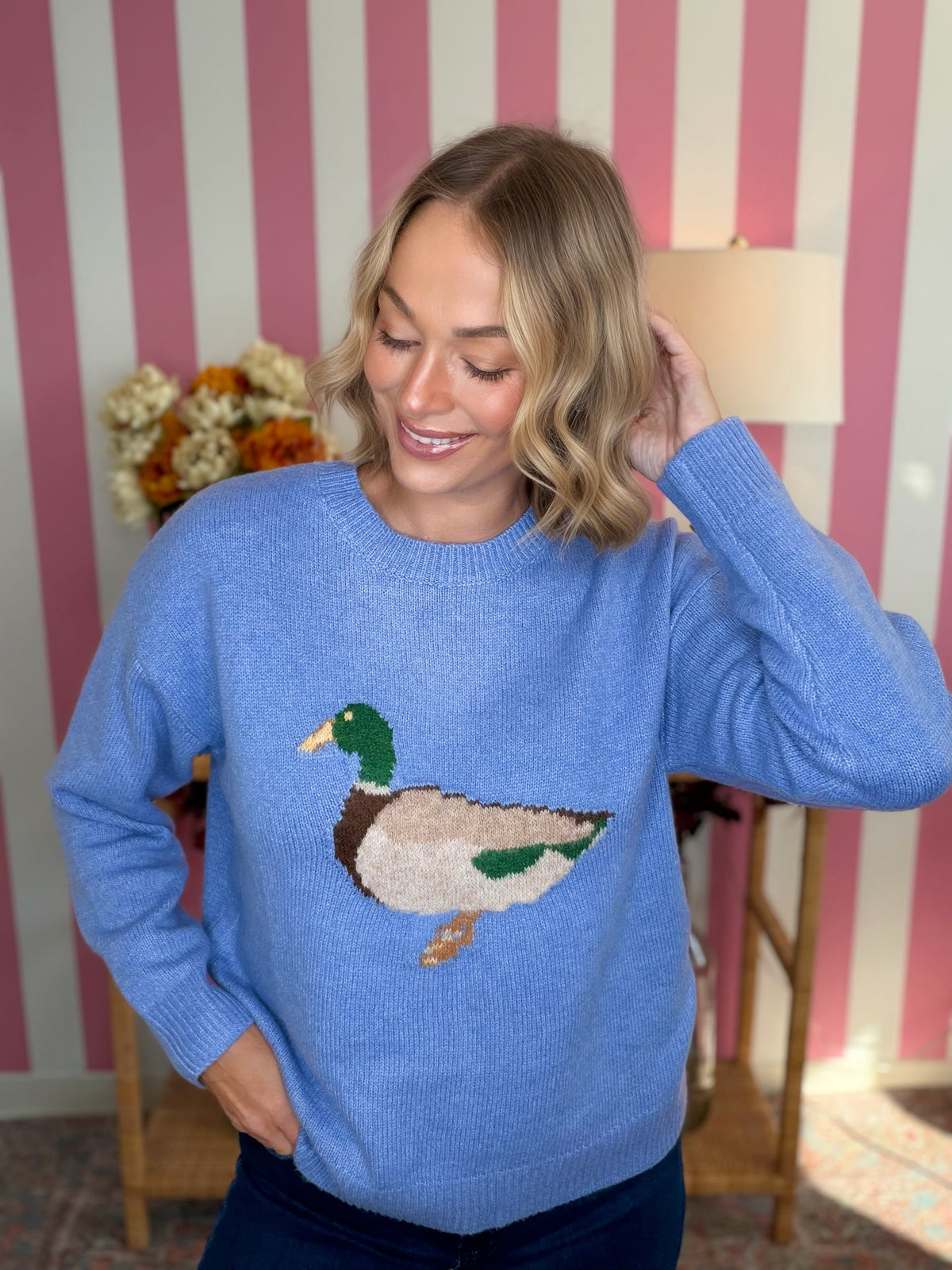 A woman wearing a light blue knit sweater with a detailed mallard duck motif on the chest, posed in front of a pink and white striped wall.
