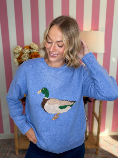 A woman wearing a light blue knit sweater with a detailed mallard duck motif on the chest, posed in front of a pink and white striped wall.