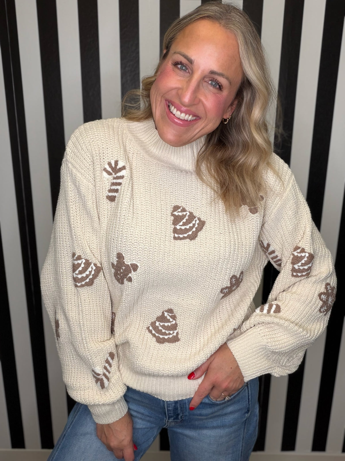 A smiling blonde woman wearing a cream-colored knit sweater with brown cupcake motifs, paired with blue jeans, standing in front of a black and white striped background.