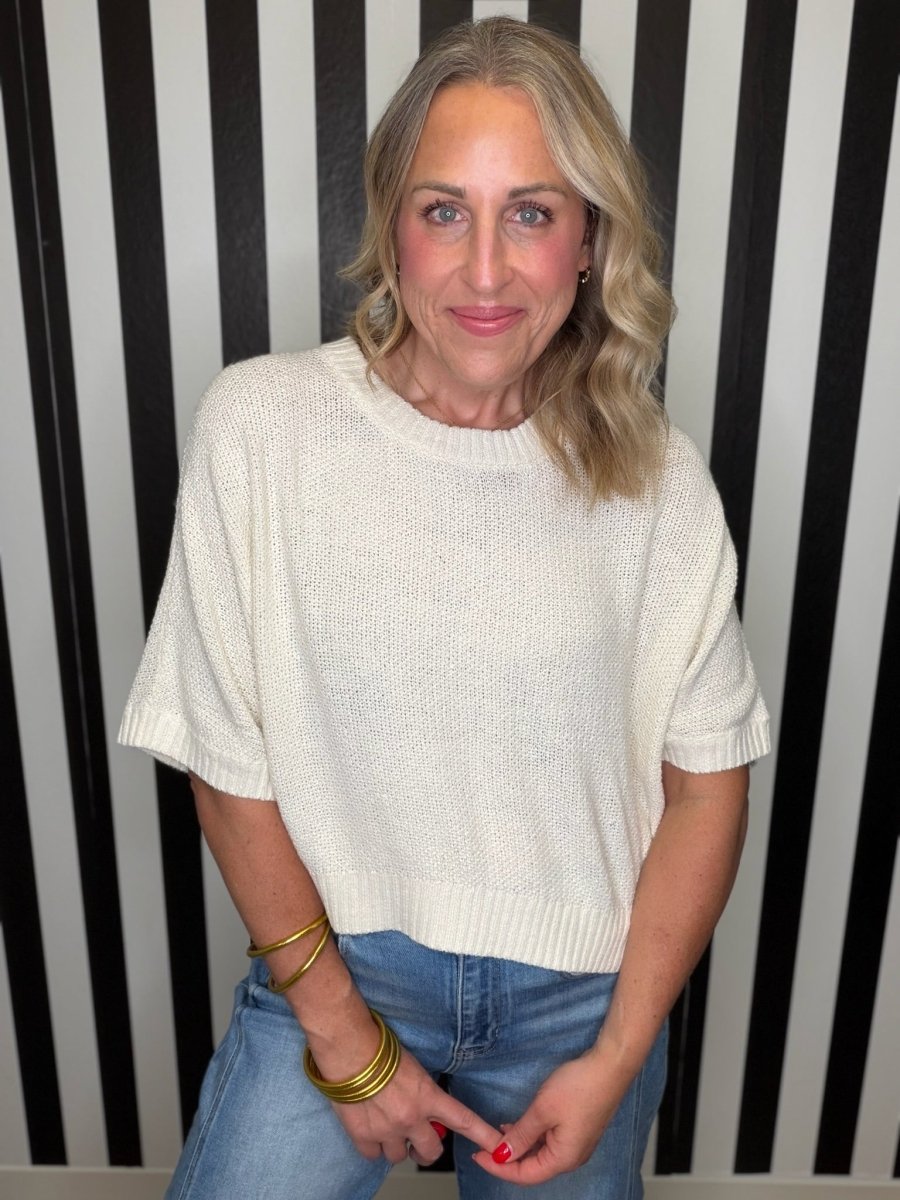 A woman with blonde hair wearing a cream-colored knit short-sleeve sweater and blue jeans, standing in front of a black and white striped wall.