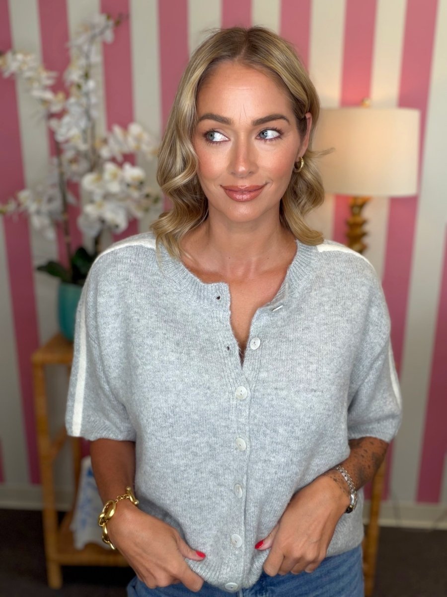 A woman wearing a light gray short-sleeve button-up cardigan with white shoulder stripes, posed in front of a pink and white striped wall.