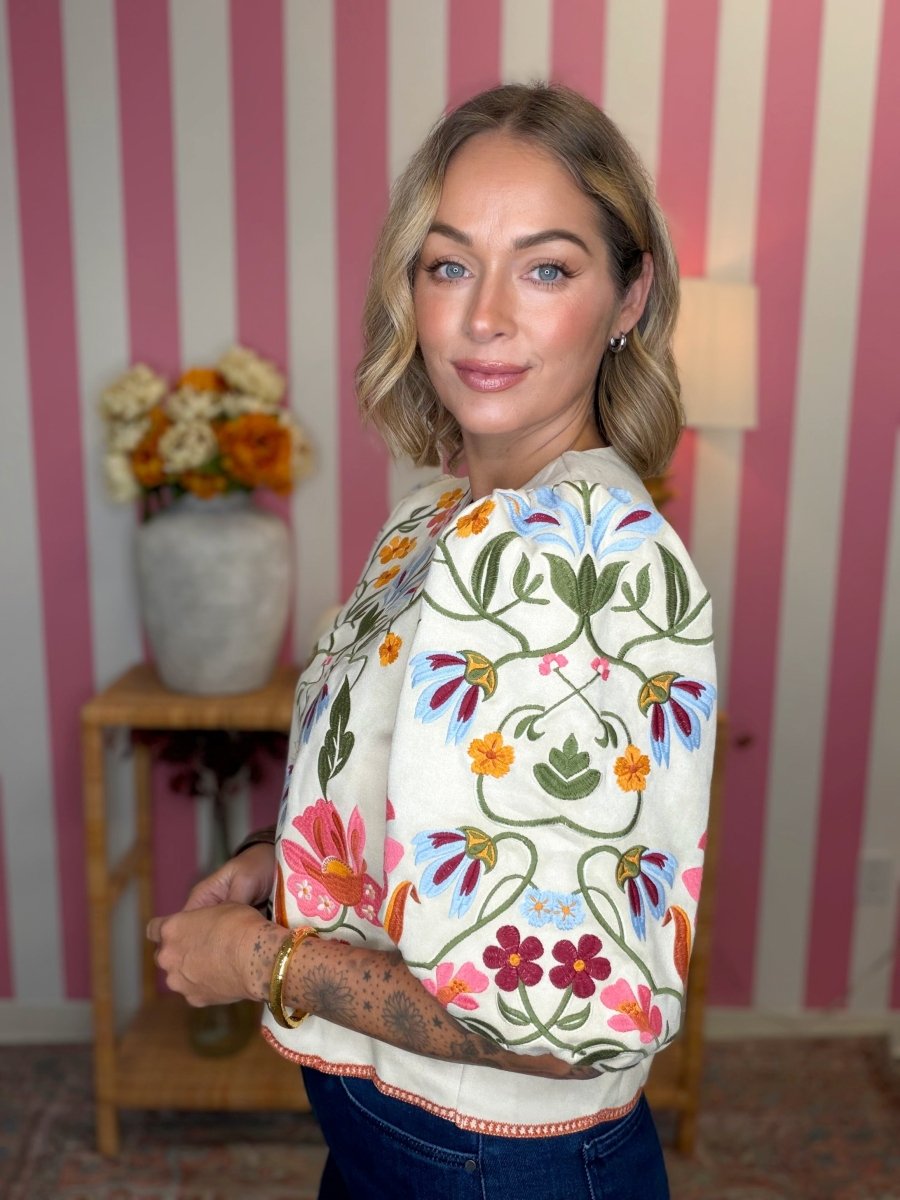 A woman with blonde wavy hair models a cream-colored short-sleeve top featuring colorful floral embroidery in pink, blue, green, and orange, paired with dark blue jeans, set against a pink and white striped wall background.