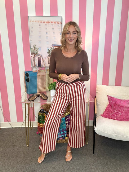 A woman stands in a room with pink and white striped wallpaper, wearing a brown long-sleeve top and red-and-white vertically striped wide-leg pants. She is posing next to a glass side table with a blue phone case and a white armchair with a pink pillow.