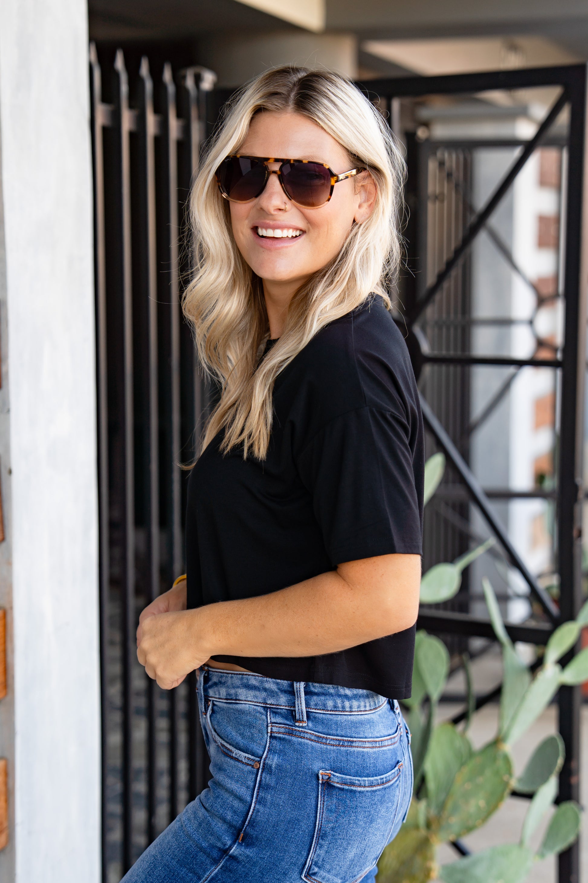 Woman wearing sunglasses and a black top with blue jeans, standing indoors.