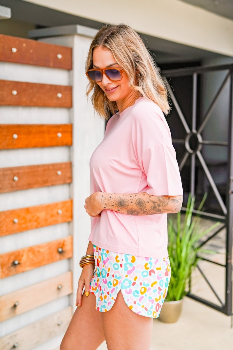 Woman wearing a pink top and colorful shorts standing in front of a wooden wall.