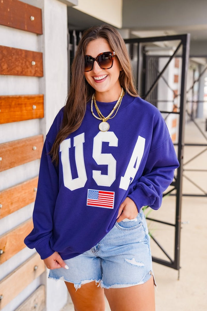 Woman wearing a blue sweatshirt with 'USA' and an American flag, standing against a wooden wall.