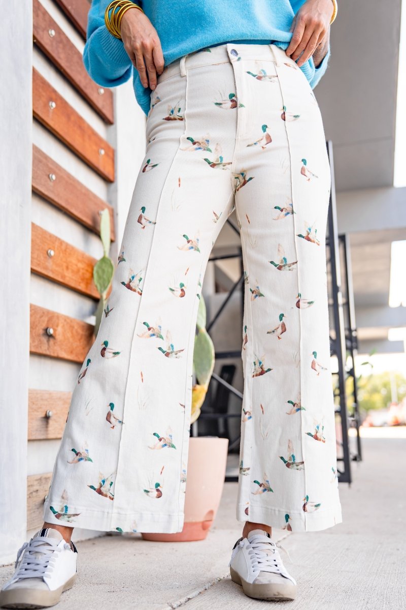 A person wearing off-white wide-leg pants with a colorful duck print, styled with a bright blue top and white sneakers in an outdoor setting.
