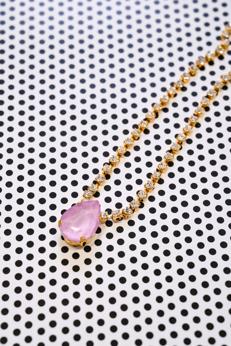 A gold chain necklace with a teardrop-shaped pink gemstone pendant, displayed diagonally on a white background with a black polka dot pattern.