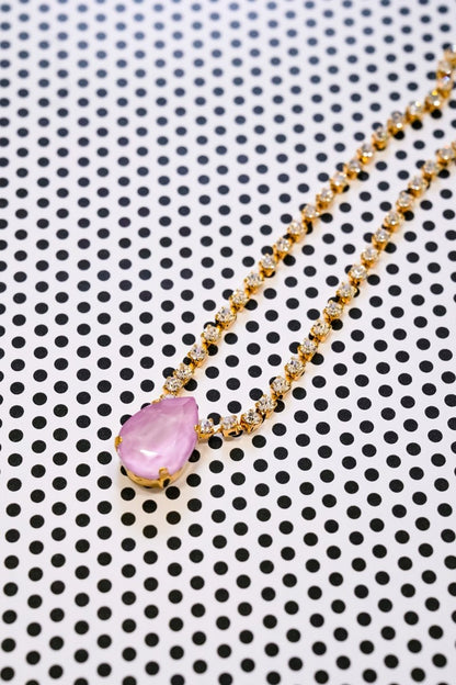 A gold chain necklace with a teardrop-shaped pink gemstone pendant, displayed diagonally on a white background with a black polka dot pattern.