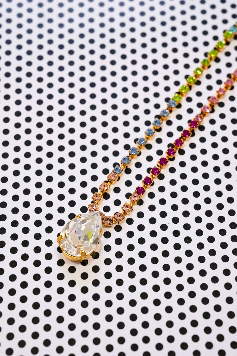 A gold-tone necklace with a large teardrop-shaped clear crystal pendant and a beaded chain featuring multicolored rhinestones, displayed on a black and white polka dot background.