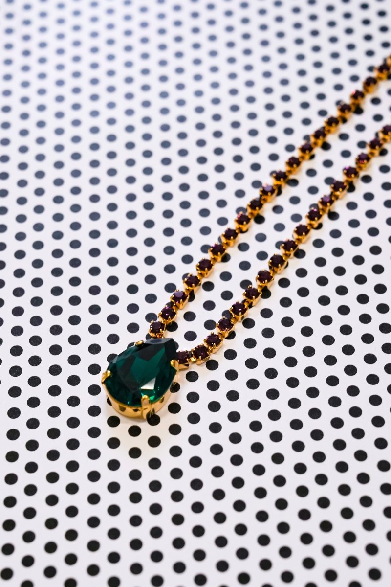 A gold necklace with a large green marquise-cut gemstone pendant and smaller gold and dark brown beads along the chain, displayed on a white background with a black polka dot pattern.